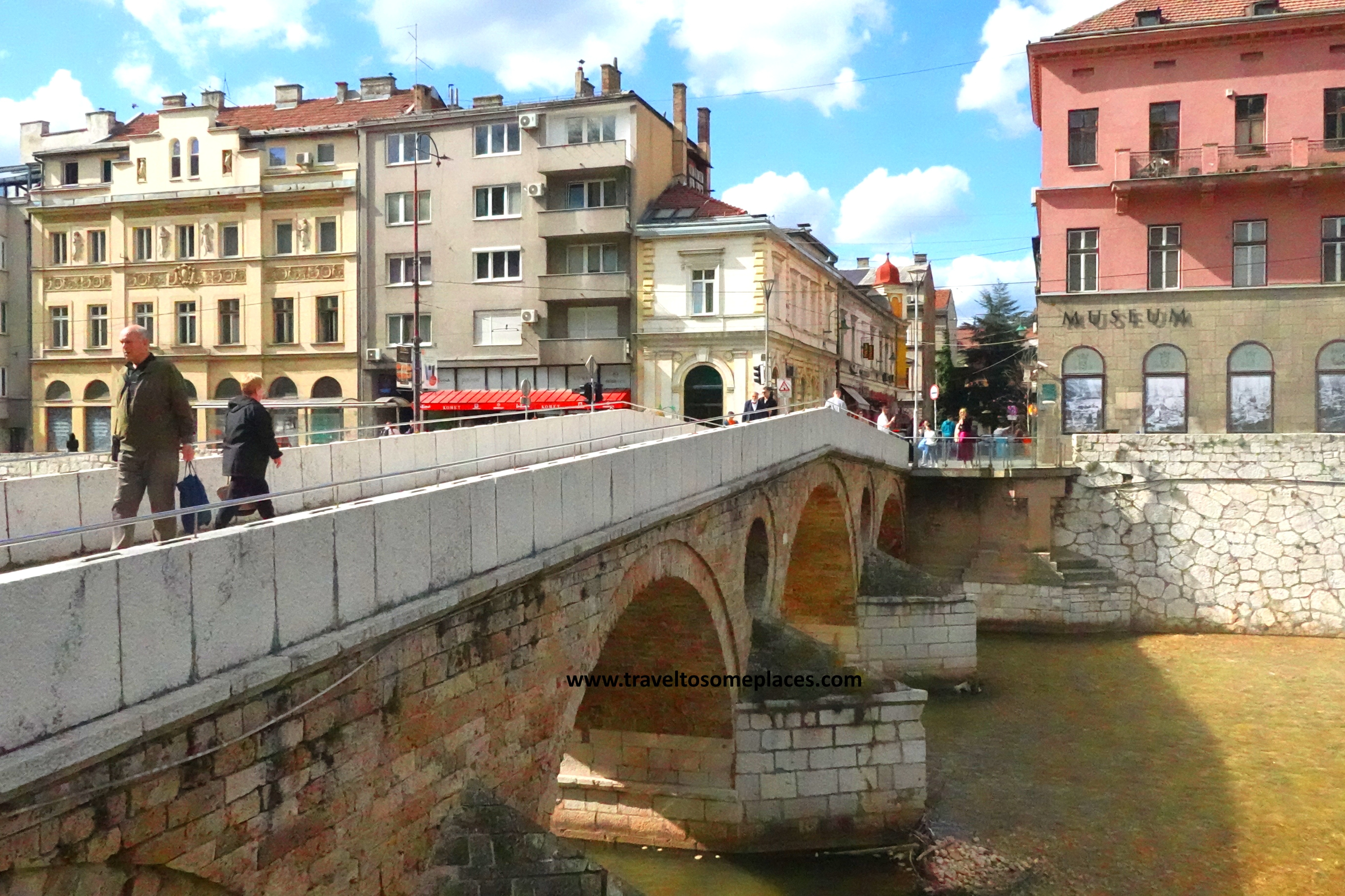 The Latin Bridge is a stone bridge in Sarajevo, Bosnia. Two people walk across the bridge which sits over a river, across we see colorful buildings.